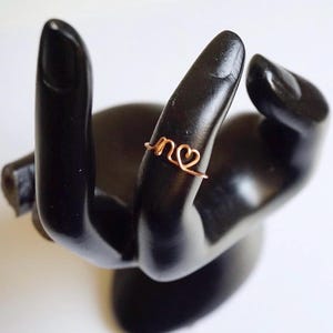 May include: A copper wire ring featuring the letters "n" and a heart design. The ring is presented on a black hand sculpture. This delicate ring is a piece of jewellery.