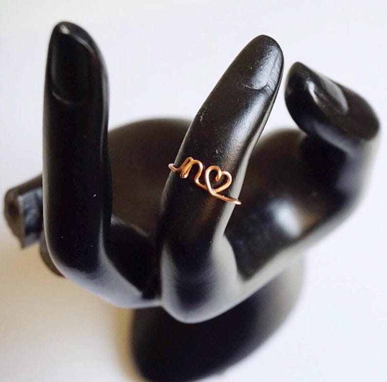 Peut inclure: Une bague en fil de cuivre de couleur cuivr&eacute;e avec les lettres "n" et un c&oelig;ur. La bague est pr&eacute;sent&eacute;e sur une sculpture noire en forme de main. Un bijou d&eacute;licat.