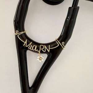 Puede incluir: Un estetoscopio negro con una etiqueta con el nombre de alambre de oro que dice "Mia RN". Un pequeño dije de flor de oro y perla cuelga debajo del nombre. El estetoscopio está sobre un fondo blanco.