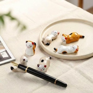 Op de afbeelding: Een verzameling keramische kattenbeeldjes in verschillende poses, waaronder staand, liggend en opgerold. De katten zijn wit, oranje en bruin met zwarte accenten. Een zwarte vulpen rust op een katvormige pennenhouder. De beeldjes staan op een beige bord.
