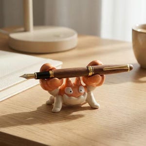 May include: A wooden fountain pen with gold detailing is held by an orange, white, and tan crab-shaped pen holder. A white notebook and a beige mug are in the background on a wooden surface.