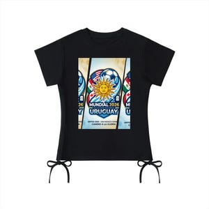 Peut inclure: T-shirt noir avec un graphisme coloré représentant un ballon de football, un soleil et le texte "Mundial 2026 Uruguay". Le t-shirt a des manches courtes et des liens décoratifs en bas.