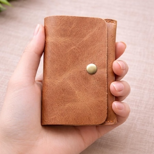 May include: A small, brown leather wallet with a button clasp. The wallet is held in a hand, highlighting its compact size and textured surface. The leather has a distressed appearance, adding to its rustic charm.