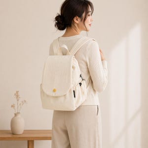 May include: Cream-colored backpack with a rounded flap and a gold-colored button closure. The backpack has adjustable straps and side pockets. The bag is made of a textured fabric and is being worn by a person.
