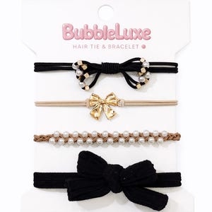 May include: A set of hair ties and bracelets on a white card. The set includes black, tan, and brown elastic bands with decorative elements such as bows and faux pearls. The brand name "BubbleLuxe" is printed in pink.
