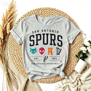 May include: A light gray t-shirt featuring the text "SAN ANTONIO SPURS" in black block letters. Below the text are colorful graphics of a fox, alien, castle, and harp. The shirt also includes the text "EST. 1973 SAN ANTONIO BASKETBALL".
