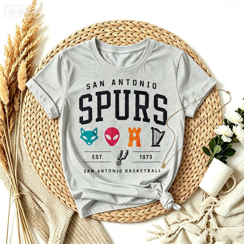 May include: A light gray t-shirt featuring the text "SAN ANTONIO SPURS" in black block letters. Below the text are colorful graphics of a fox, alien, castle, and harp. The shirt also includes the text "EST. 1973 SAN ANTONIO BASKETBALL".