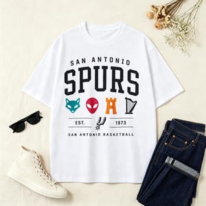 May include: White t-shirt with the text "SAN ANTONIO SPURS" in black block letters. Below the text are colorful graphics of a fox, alien, castle, and harp. The shirt also includes the text "EST. 1973 SAN ANTONIO BASKETBALL".
