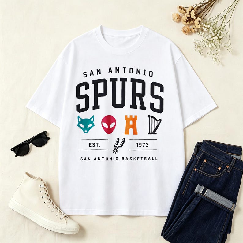 May include: White t-shirt with the text "SAN ANTONIO SPURS" in black block letters. Below the text are colorful graphics of a fox, alien, castle, and harp. The shirt also includes the text "EST. 1973 SAN ANTONIO BASKETBALL".