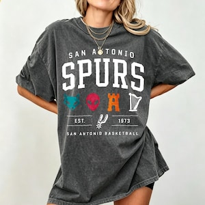 May include: Dark gray oversized t-shirt featuring the text "SAN ANTONIO SPURS" in white block letters. Below the text are various colorful graphics and the text "EST. 1973 SAN ANTONIO BASKETBALL".