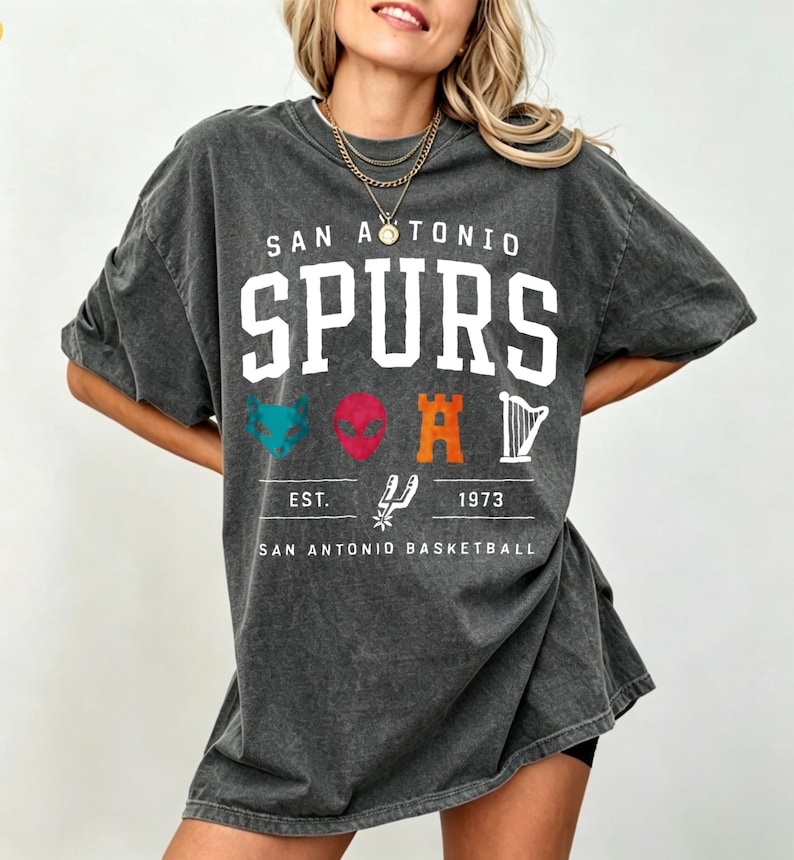 May include: Dark gray oversized t-shirt featuring the text "SAN ANTONIO SPURS" in white block letters. Below the text are various colorful graphics and the text "EST. 1973 SAN ANTONIO BASKETBALL".