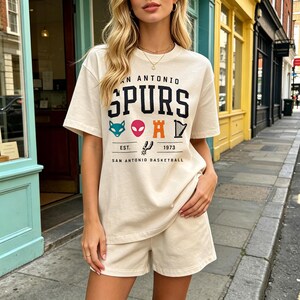 May include: Cream-colored t-shirt and shorts set. The t-shirt features the text "SAN ANTONIO SPURS" in black, with colorful graphics and the text "EST. 1973 SAN ANTONIO BASKETBALL". The shorts match the shirt.
