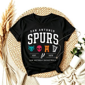 May include: Black San Antonio Spurs t-shirt with a white logo. The logo includes the team name, the text "EST. 1973", and various graphics. The shirt is displayed on a woven mat with decorative elements.