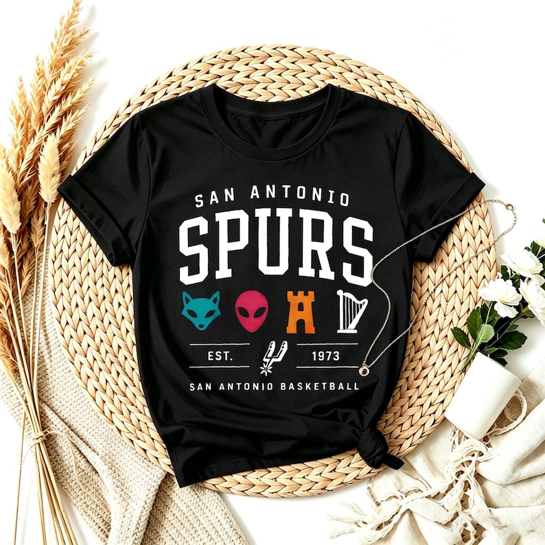 May include: Black San Antonio Spurs t-shirt with a white logo. The logo includes the team name, the text "EST. 1973", and various graphics. The shirt is displayed on a woven mat with decorative elements.