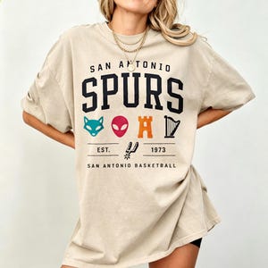 May include: Beige t-shirt with the text "SAN ANTONIO SPURS" in black block letters. Below the text are various colorful graphics and the text "EST. 1973 SAN ANTONIO BASKETBALL". The shirt is a casual, oversized style.