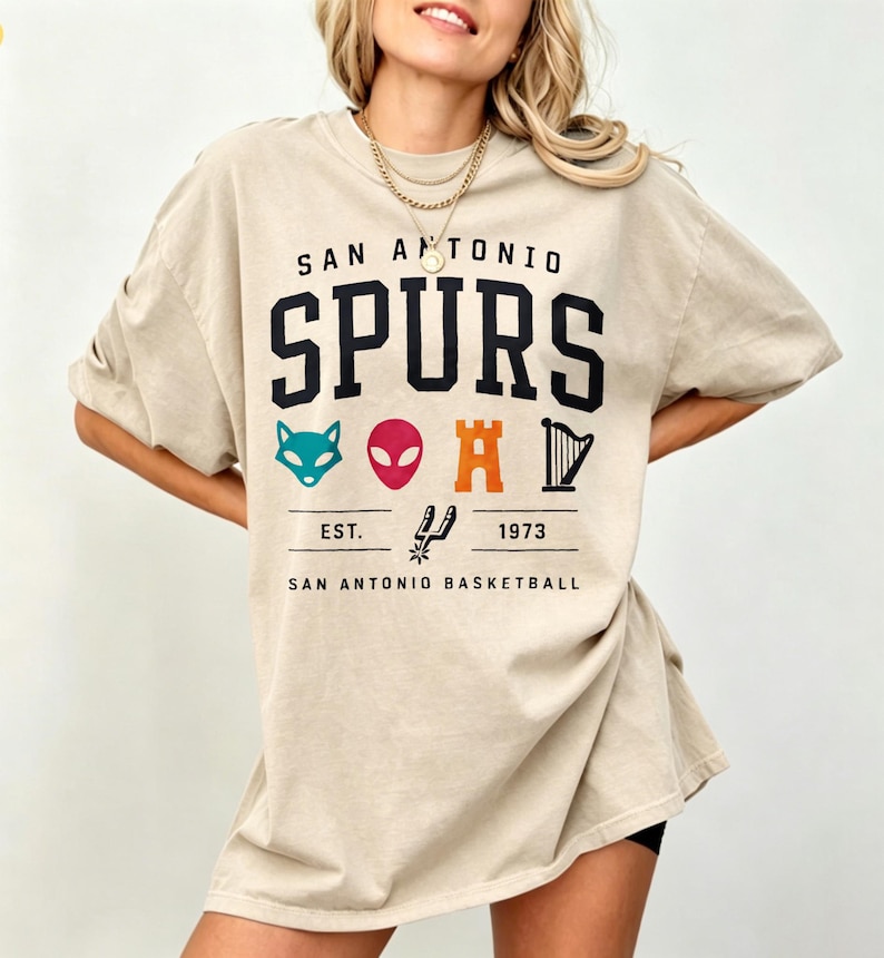 May include: Beige t-shirt with the text "SAN ANTONIO SPURS" in black block letters. Below the text are various colorful graphics and the text "EST. 1973 SAN ANTONIO BASKETBALL". The shirt is a casual, oversized style.
