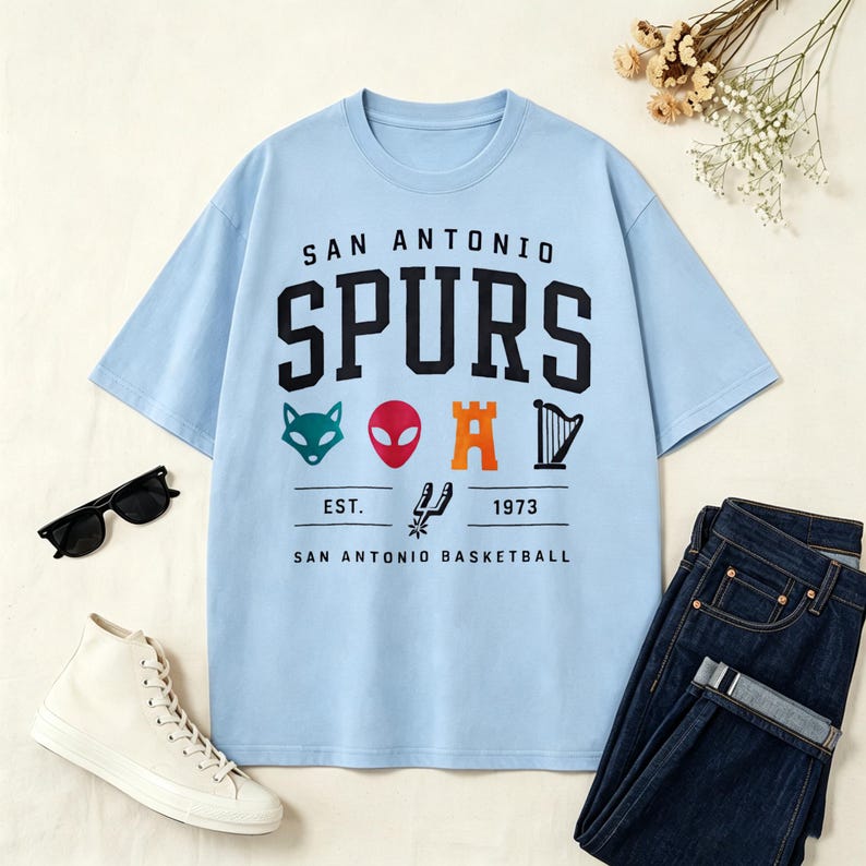 May include: Light blue t-shirt featuring the text "SAN ANTONIO SPURS" in black, with various graphics below. The shirt includes the text "EST. 1973 SAN ANTONIO BASKETBALL". The shirt is paired with dark blue jeans, white sneakers, and black sunglasses.