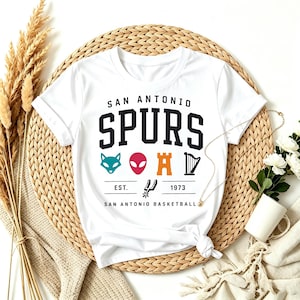 May include: White t-shirt featuring the text "SAN ANTONIO SPURS" in black block letters. Below the text are various colorful graphics and the text "EST. 1973 SAN ANTONIO BASKETBALL". The shirt is displayed on a woven mat.