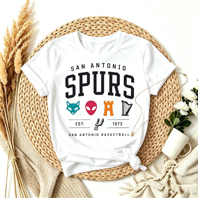 May include: White t-shirt featuring the text "SAN ANTONIO SPURS" in black block letters. Below the text are various colorful graphics and the text "EST. 1973 SAN ANTONIO BASKETBALL". The shirt is displayed on a woven mat.