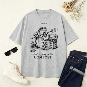 Op de afbeelding: Lichtgrijs t-shirt met een cartoon kikker die een hooivork vasthoudt, staand voor een houten kist. De tekst op het shirt luidt: "That's it. You're going in the COMPOST."