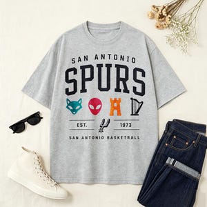 May include: A gray t-shirt with the words "SAN ANTONIO SPURS" in black block letters. Below the text are colorful graphics and the text "EST. 1973 SAN ANTONIO BASKETBALL". The shirt is paired with blue jeans and white sneakers.