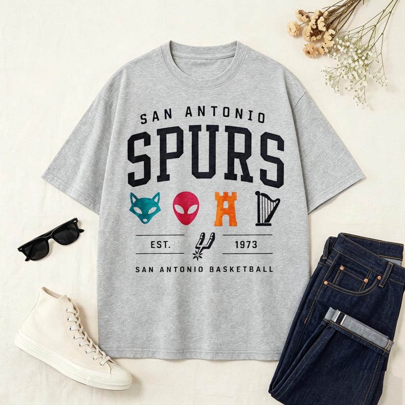 May include: A gray t-shirt with the words "SAN ANTONIO SPURS" in black block letters. Below the text are colorful graphics and the text "EST. 1973 SAN ANTONIO BASKETBALL". The shirt is paired with blue jeans and white sneakers.