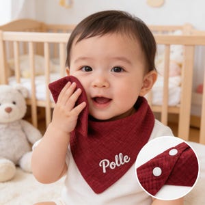 Pode incluir: Babete de bebé bordô com o nome "Polle" bordado em branco. O babete é triangular e tem dois fechos de pressão brancos. O bebé segura uma toalha de rosto bordô a condizer.