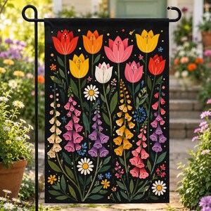 May include: A black garden flag featuring a colorful floral design. The design includes tulips, foxgloves, and daisies in shades of red, orange, pink, yellow, white, and purple. The flag is displayed on a black metal stand in a garden setting.