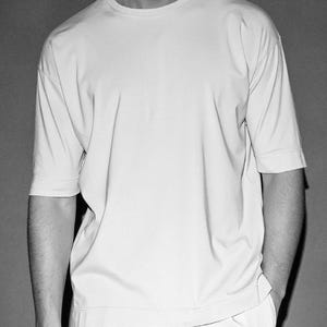 Op de afbeelding: Een zwart-wit afbeelding van een lichtgekleurd, kort mouw t-shirt met een oversized pasvorm. Het shirt heeft een eenvoudig ontwerp en wordt gedragen door een persoon met de handen in de zakken. De stof lijkt glad te zijn.