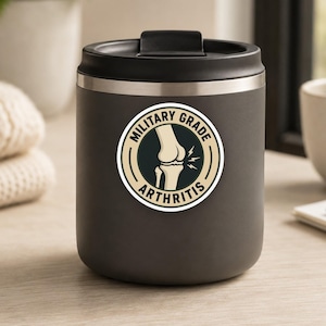 May include: A matte charcoal gray insulated tumbler with a circular sticker featuring a beige background, black text reading "MILITARY GRADE ARTHRITIS," and a central illustration of a human knee joint with small lightning bolt icons.