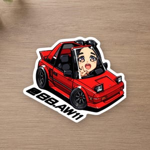 May include: A die-cut sticker featuring a red sports car with pop-up headlights and a chibi-style character in the driver seat. The character has dark hair and is making a peace sign. The text 88.AW11 is printed in black on the white border.