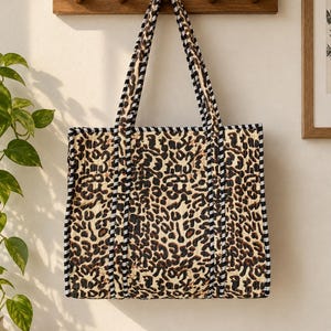 May include: A rectangular tote bag featuring a tan and black leopard print pattern. The bag is constructed with quilted fabric and is finished with black and white striped piping along the edges and the two shoulder straps.