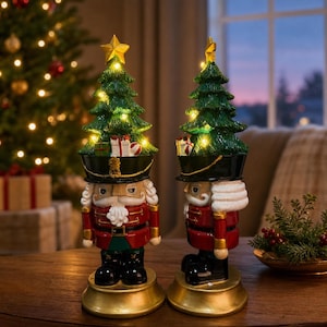 Könnte beinhalten: Zwei Nussknacker-Figuren auf goldenen runden Sockeln. Jeder Nussknacker trägt einen schwarzen Hut, auf dem ein grüner Weihnachtsbaum mit kleinen leuchtenden Lichtern und Miniatur-Geschenkboxen sitzt. Sie tragen rote Jacken mit Goldakzenten.