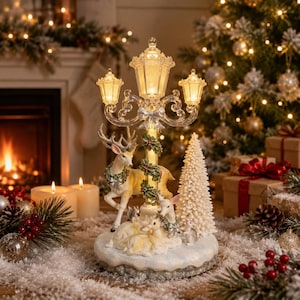 Può includere: Una lampada da tavolo decorativa con un design a lampione centrale con tre lanterne illuminate, decorata con un fiocco argentato e vegetazione. Un cervo e due cerbiatti si trovano alla base vicino a un albero di Natale bianco testurizzato.