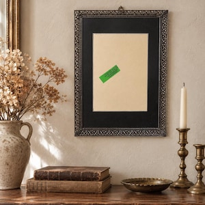 May include: A rectangular wall frame with a dark, ornate, filigree-style metal border and a black mat surrounding a central beige rectangular insert. A small, bright green rectangular sticker with illegible text is affixed to the beige centre.