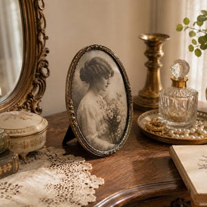 Puede incluir: Un marco de fotos ovalado con un borde metálico oscuro y ornamentado que contiene un retrato en blanco y negro de una persona sosteniendo un ramo de flores. El marco presenta detalles en relieve intrincados en el borde y se mantiene en pie.
