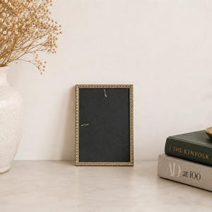 Puede incluir: Un marco de fotos rectangular con un borde fino de color dorado texturizado y un inserto negro sólido. El marco se mantiene en posición vertical, presenta un patrón similar a una cuerda en los bordes y está situado entre un jarrón blanco y libros.