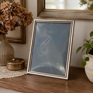 May include: A rectangular picture frame with a metallic silver finish and a thin, raised border. The frame contains a clear protective sheet with a faint, white outline of a sock shape printed on the surface, set against a dark blue background.