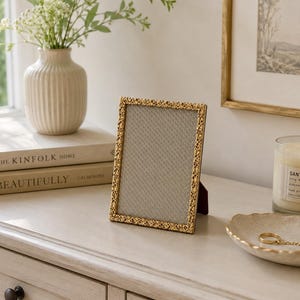 Puede incluir: Un marco de fotos rectangular con un borde metálico dorado texturizado que presenta un intrincado patrón floral en relieve. El marco contiene una paspartú de color gris claro con textura fina y se apoya en un soporte trasero tipo caballete.