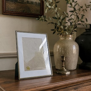 Puede incluir: Un marco de fotos rectangular con un borde metálico fino y un paspartú blanco. El marco contiene un inserto beige texturizado con un pequeño sello dorado circular en la esquina inferior izquierda. El marco está colocado sobre una superficie de madera.