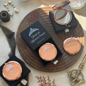 以下が含まれることがあります： ローズゴールドのコンパクトミラー。一部は開いており、一部は黒い箱に入っています。「Sweet 16」と名前が書かれています。ミラーは丸く、反射面があります。箱は木製の表面に置かれ、装飾的な要素があります。