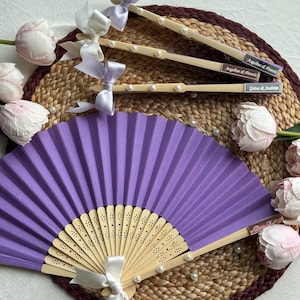 May include: A purple folding fan with a wooden handle and a white ribbon, alongside decorated chopsticks with pearls and ribbons. The chopsticks have names printed on them. The items are arranged on a woven mat with floral accents.