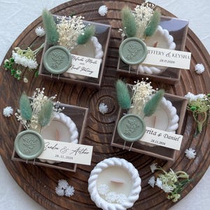 Puede incluir: Cuatro velas decorativas en cajas transparentes, cada una con un borde blanco en forma de cuerda y un pequeño arreglo floral. Cada caja tiene una etiqueta con nombre personalizado y un sello de cera verde. Las velas están dispuestas sobre una bandeja de madera.