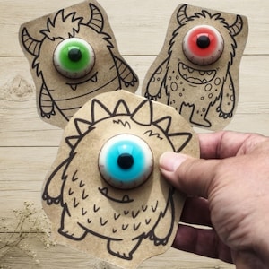 Könnte beinhalten: Drei gestanzte Karten aus Kraftpapier mit schwarzen Tuschezeichnungen von Monstern, die jeweils mit einem großen, farbigen, kugelförmigen Auge versehen sind. Die Monster haben unterschiedliche Formen, Hörner und Texturen, mit grünen, roten und blauen Augen.