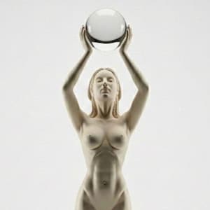 May include: A nude sculpture of a woman holding a clear glass sphere above her head. The figure is standing on a round base, with arms extended upwards. The sculpture is a light beige color.