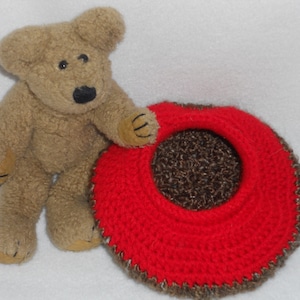 Può includere: Un orsacchiotto marrone con un cappello all'uncinetto rosso e marrone. Il cappello ha un bordo all'uncinetto marrone e una parte superiore all'uncinetto rossa.
