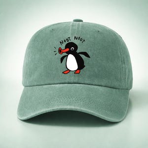 May include: A sage green baseball cap featuring a cartoon penguin with a red beak and feet, and a white belly. The penguin is surrounded by the words "NOOT NOOT" in black. The cap has a curved brim and an adjustable strap.
