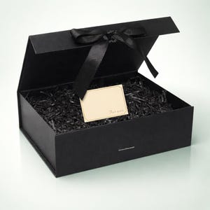 Puede incluir: Una caja de regalo negra con una cinta a juego y una pequeña tarjeta que dice "For you". La caja está abierta, revelando papel negro triturado en el interior. La caja es rectangular y tiene un cierre magnético.