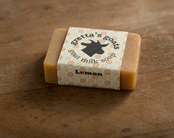 May include: A bar of goat milk soap with a label that reads "gretta's goats goat milk soap Lemon" and features a small goat illustration. The soap is wrapped in a floral pattern.