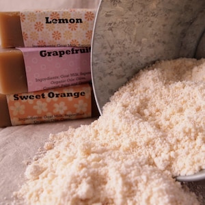May include: Three bars of goat milk soap, stacked on top of each other. The bars are labeled "Lemon", "Grapefruit", and "Sweet Orange". The soap is spilling out of a metal container.